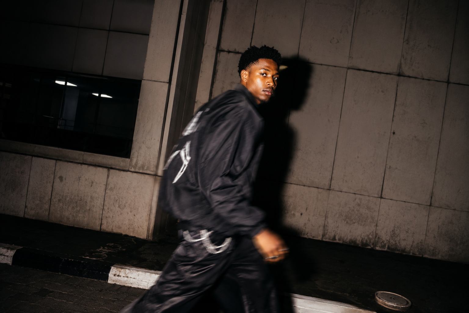A figure walking past a concrete wall at night, caught by hard on-camera flash with a bold cast shadow.